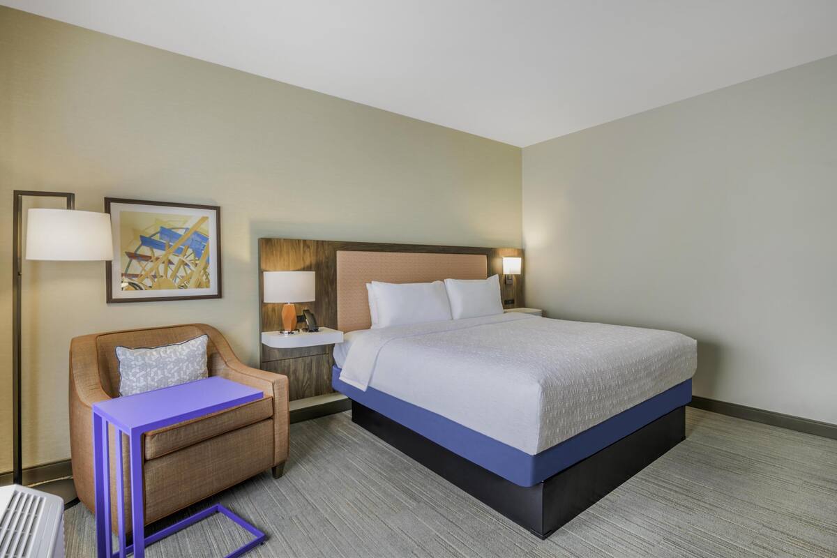 A hotel room with a bed, a chair, a small table, and a nightstand with lamps.