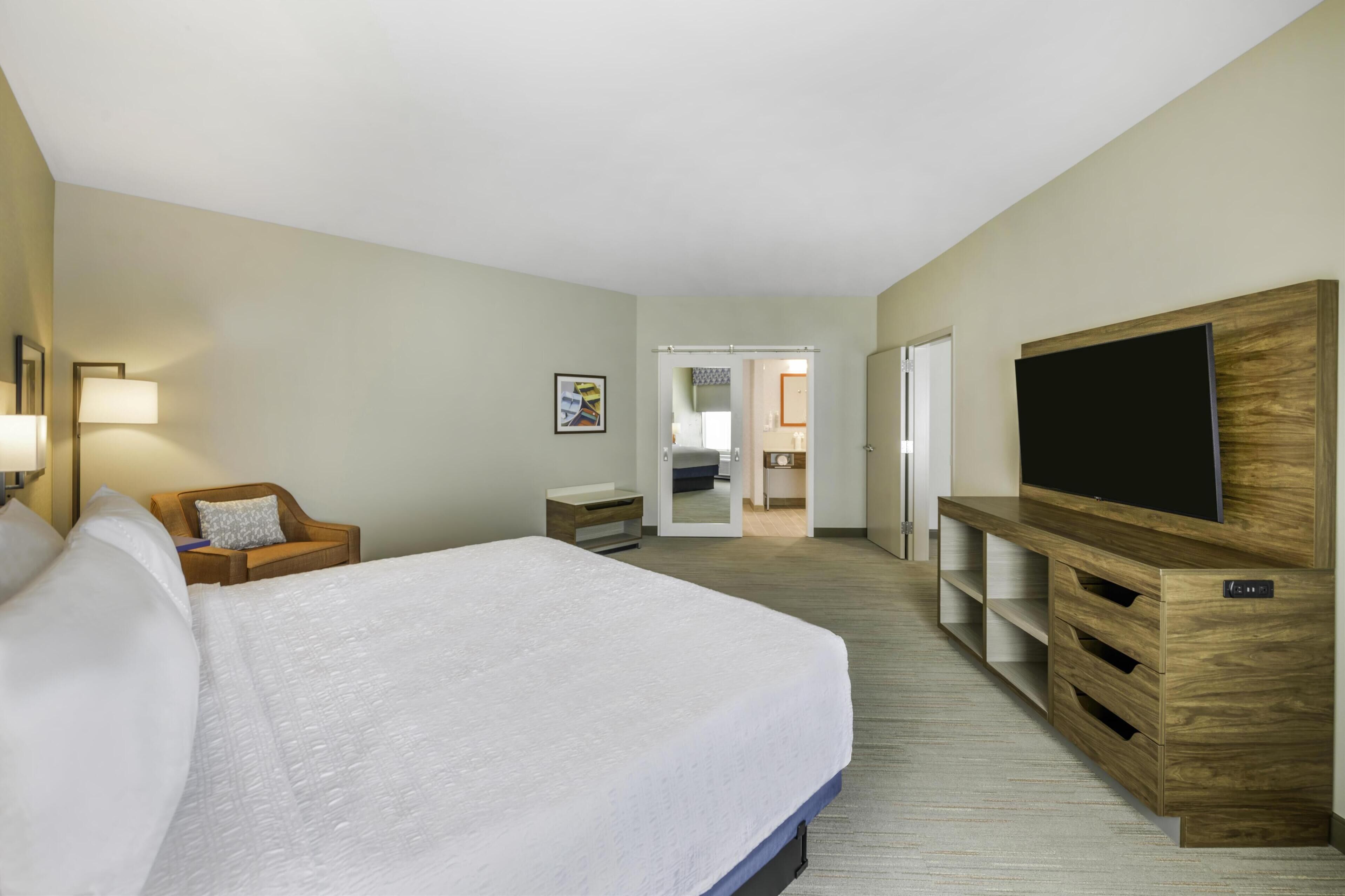 Une chambre d’hôtel avec un grand lit, un téléviseur posé sur un meuble en bois, une chaise, une table de chevet et une porte donnant sur une salle de bain.
