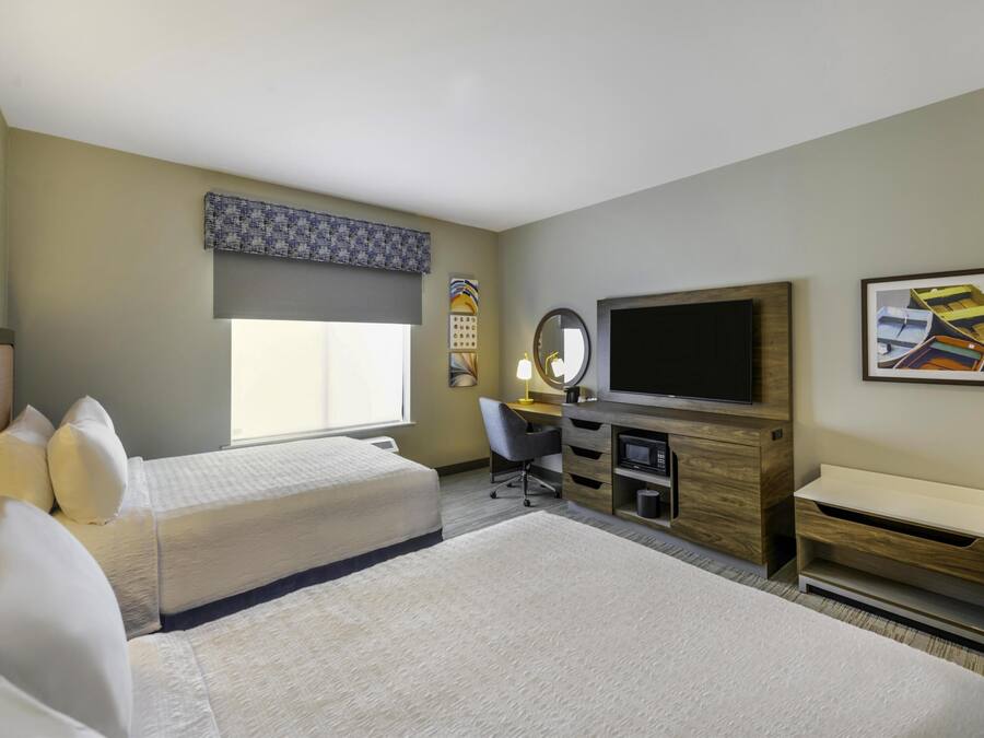 Quarto de hotel com duas camas, uma escrivaninha, televisão e uma janela grande.