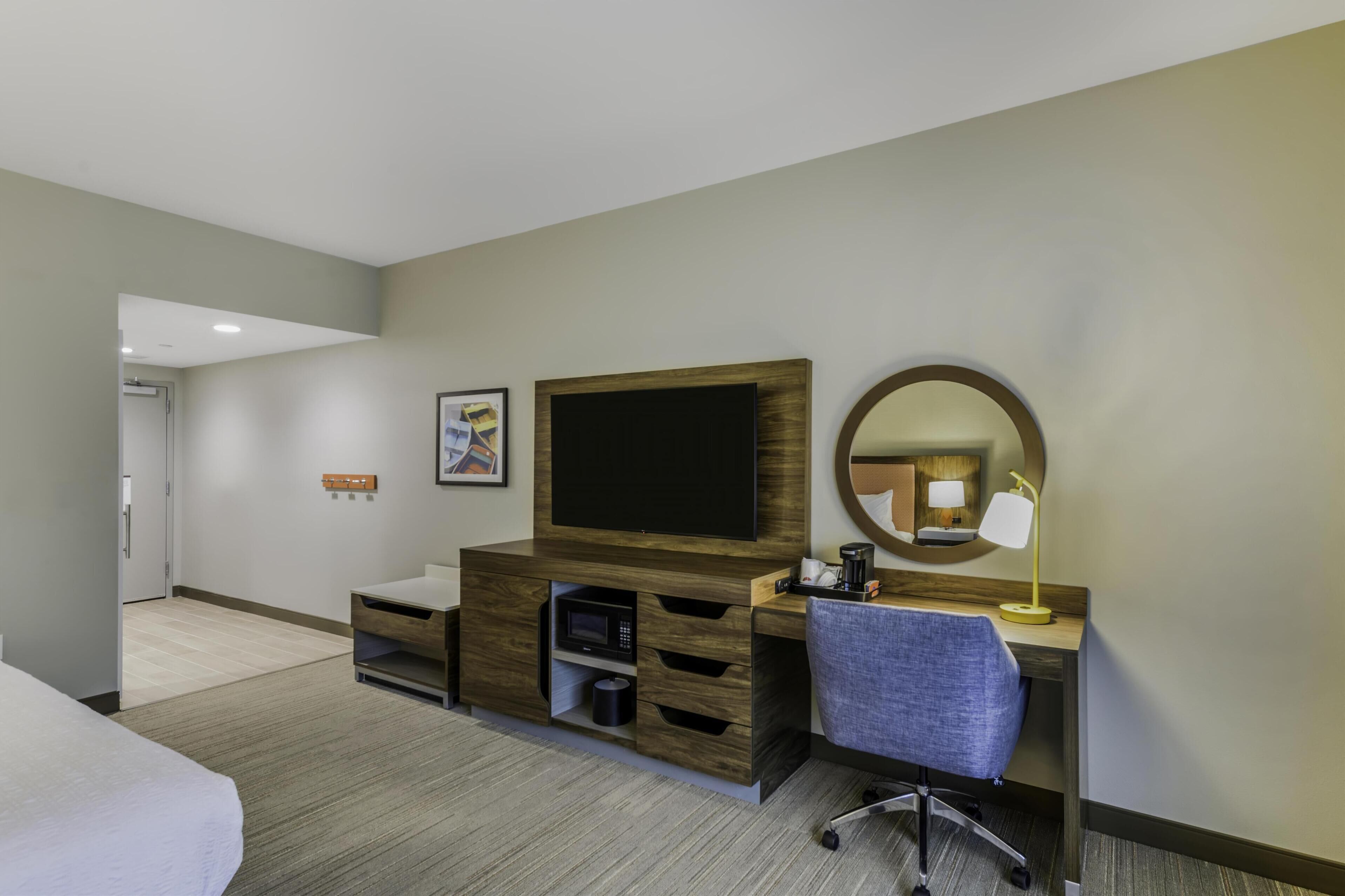 A hotel room with a bed, a desk with a computer, a flat-screen TV, a microwave, a coffee maker, and a mirror.