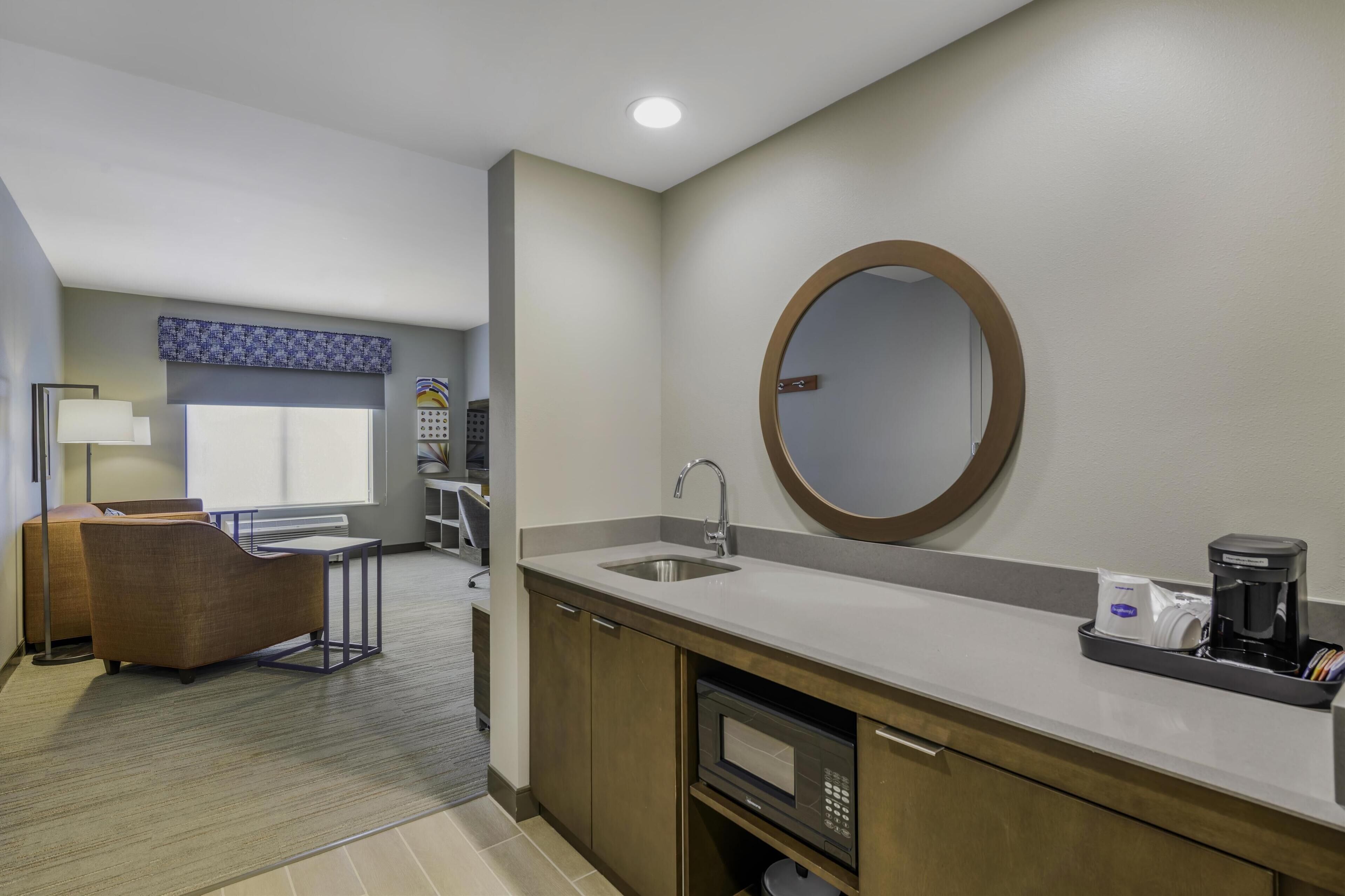 A modern hotel bathroom with a sink, mirror, and coffee maker. There is a seating area with a chair and a table in the background.