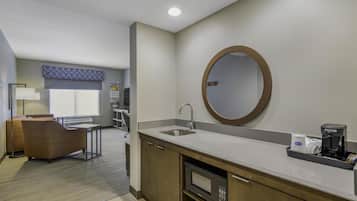 A modern hotel bathroom with a sink, mirror, and coffee maker. There is a seating area with a chair and a table in the background.