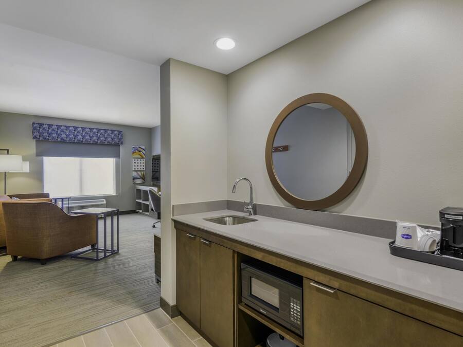 A modern hotel bathroom with a sink, mirror, and coffee maker. There is a seating area with a chair and a table in the background.