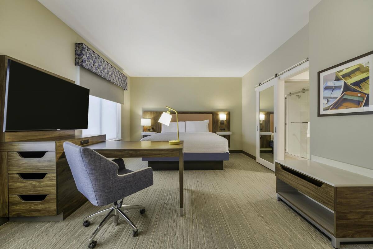 A hotel room with a bed, desk, chair, TV, lamp, wardrobe, and a mirror.
