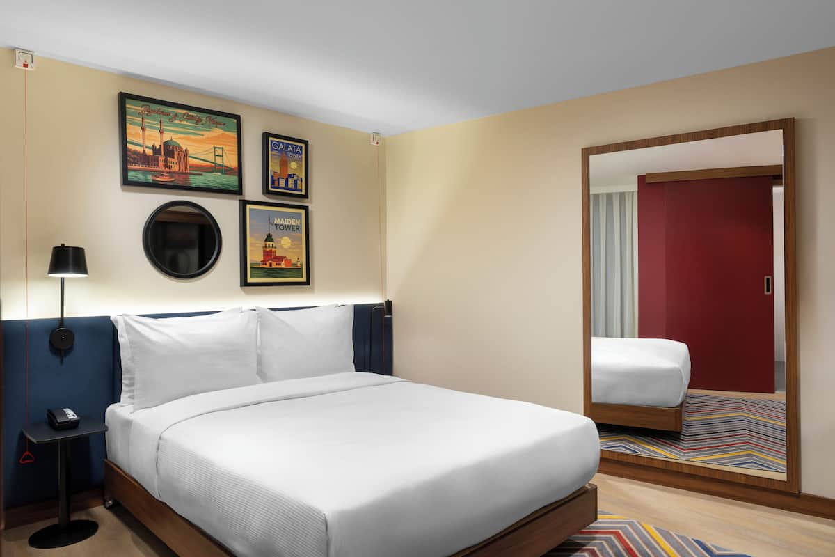 Habitación de hotel con una cama grande, una mesita de noche con lámpara y un espejo que refleja la habitación.