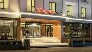 Das Gebäude eines Hampton by Hilton Hotels mit moderner Fassade, großen Fenstern und einem markanten Eingang mit Glastüren.