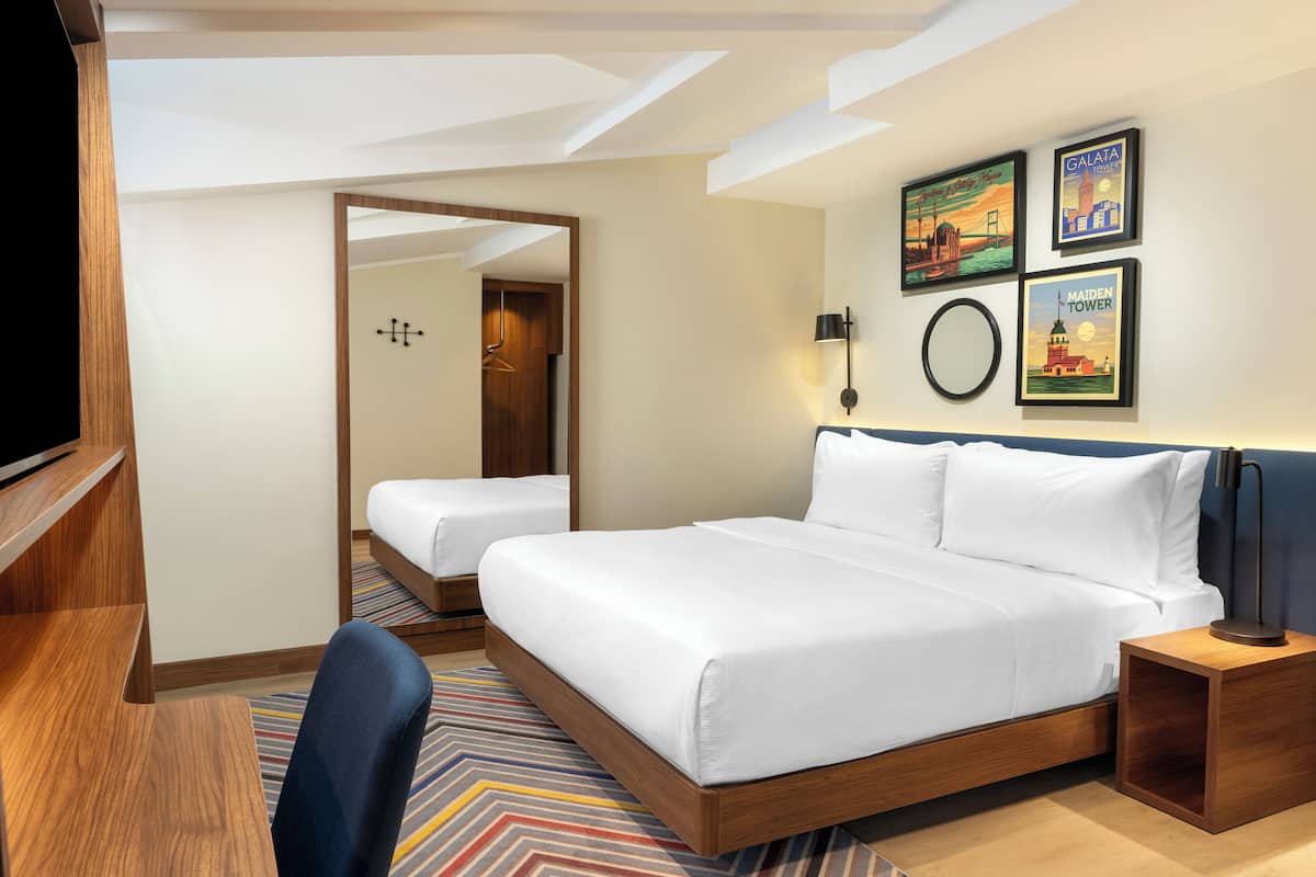 Habitación de hotel con una cama grande, una cama más pequeña, un escritorio y una silla. Hay cuadros enmarcados en la pared y un espejo.