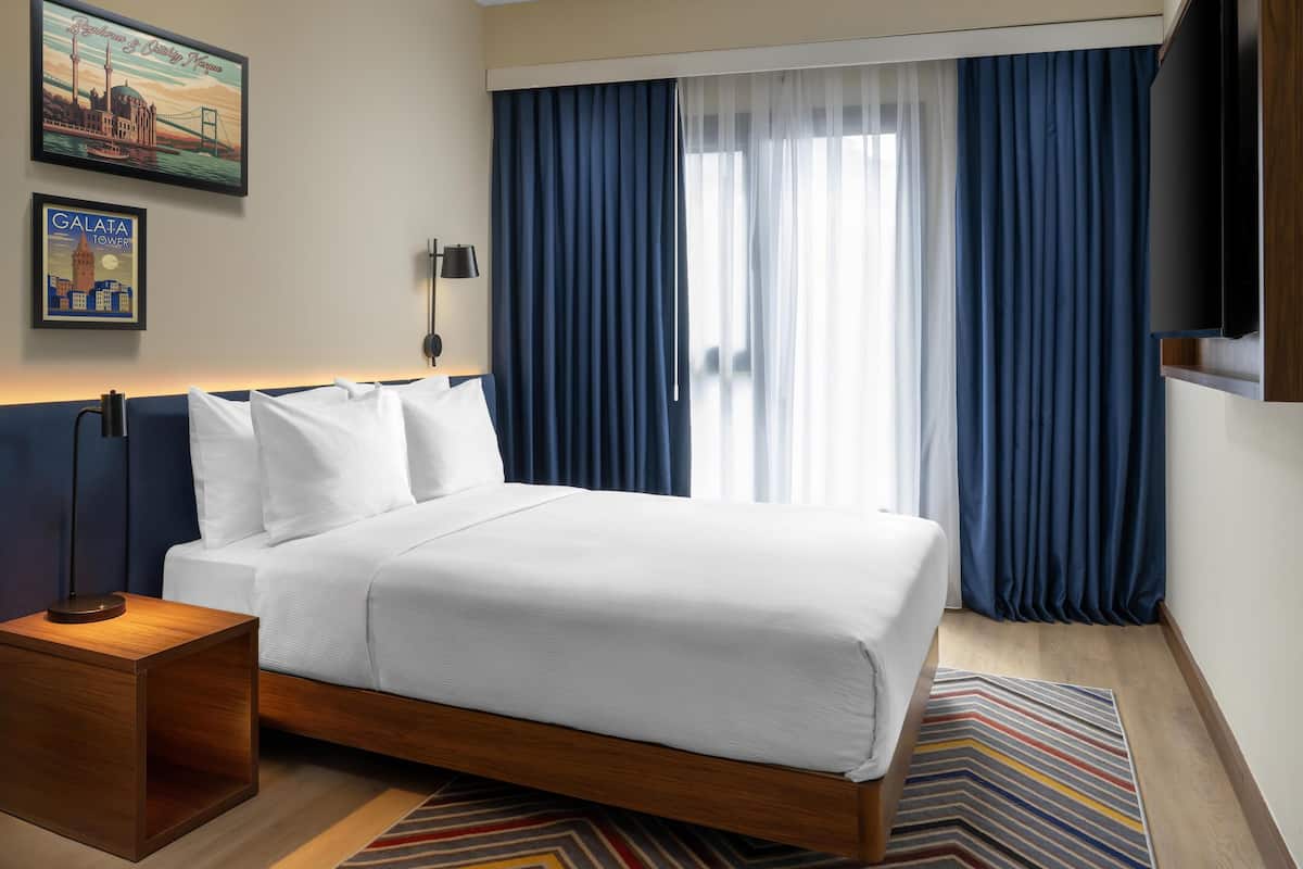 Una habitación de hotel con cama, mesita de noche, lámpara, televisor y ventana con cortinas.