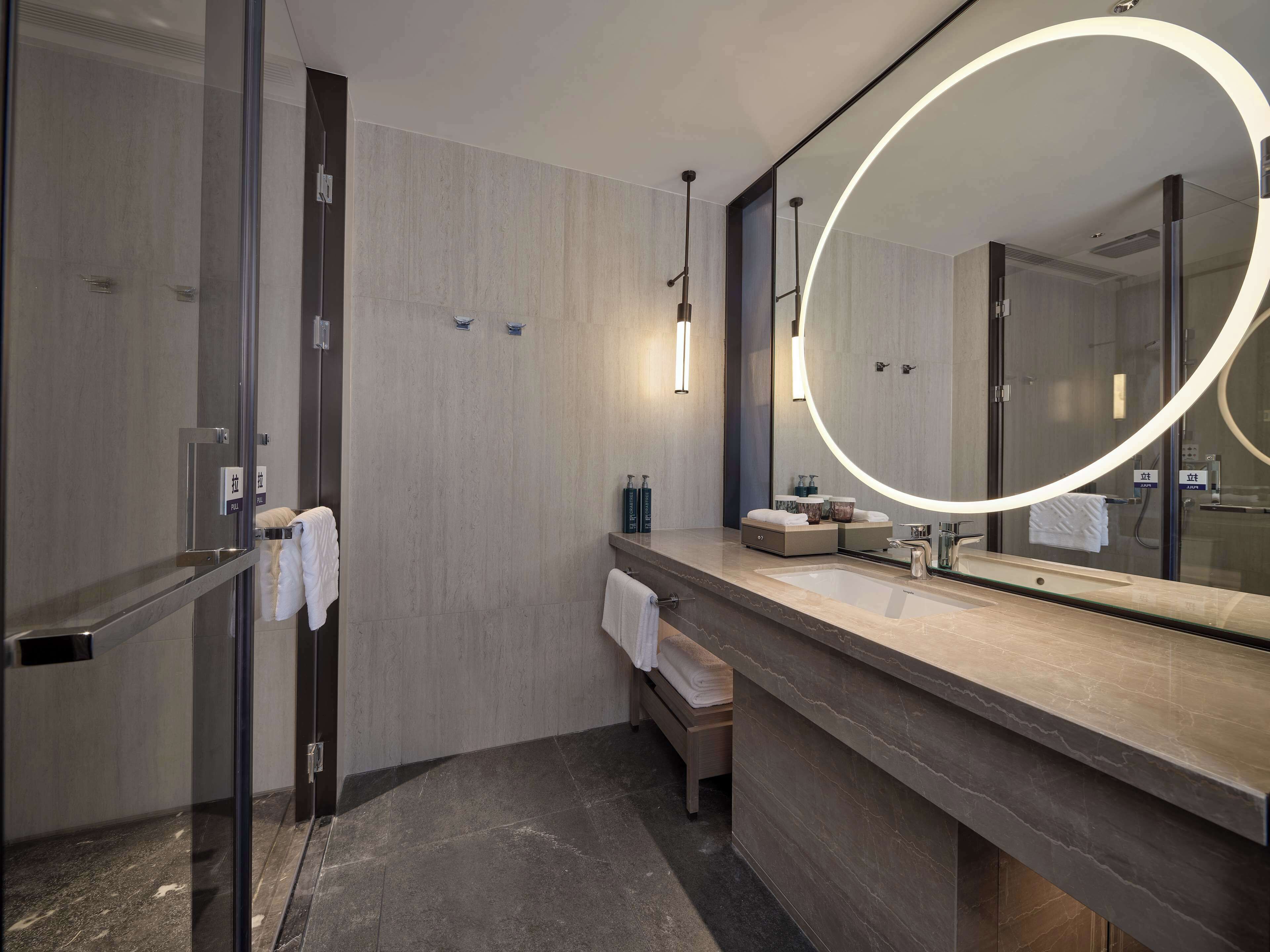Un baño moderno con cabina de ducha de vidrio, un espejo grande con luz circular y encimera de mármol.