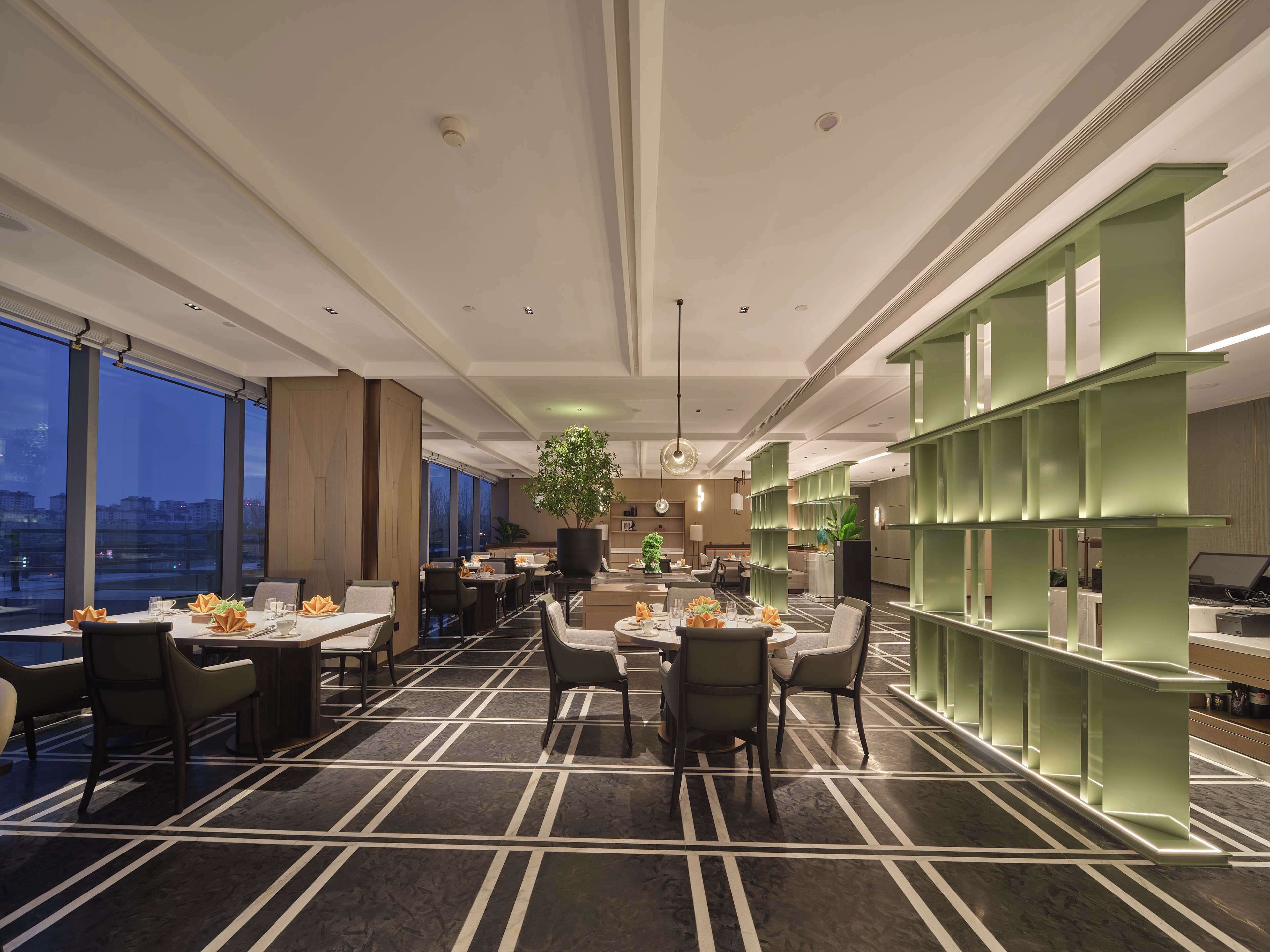 Un comedor moderno con mesas dispuestas para cuatro personas, una planta grande en maceta y vistas de la ciudad a través de amplios ventanales.
