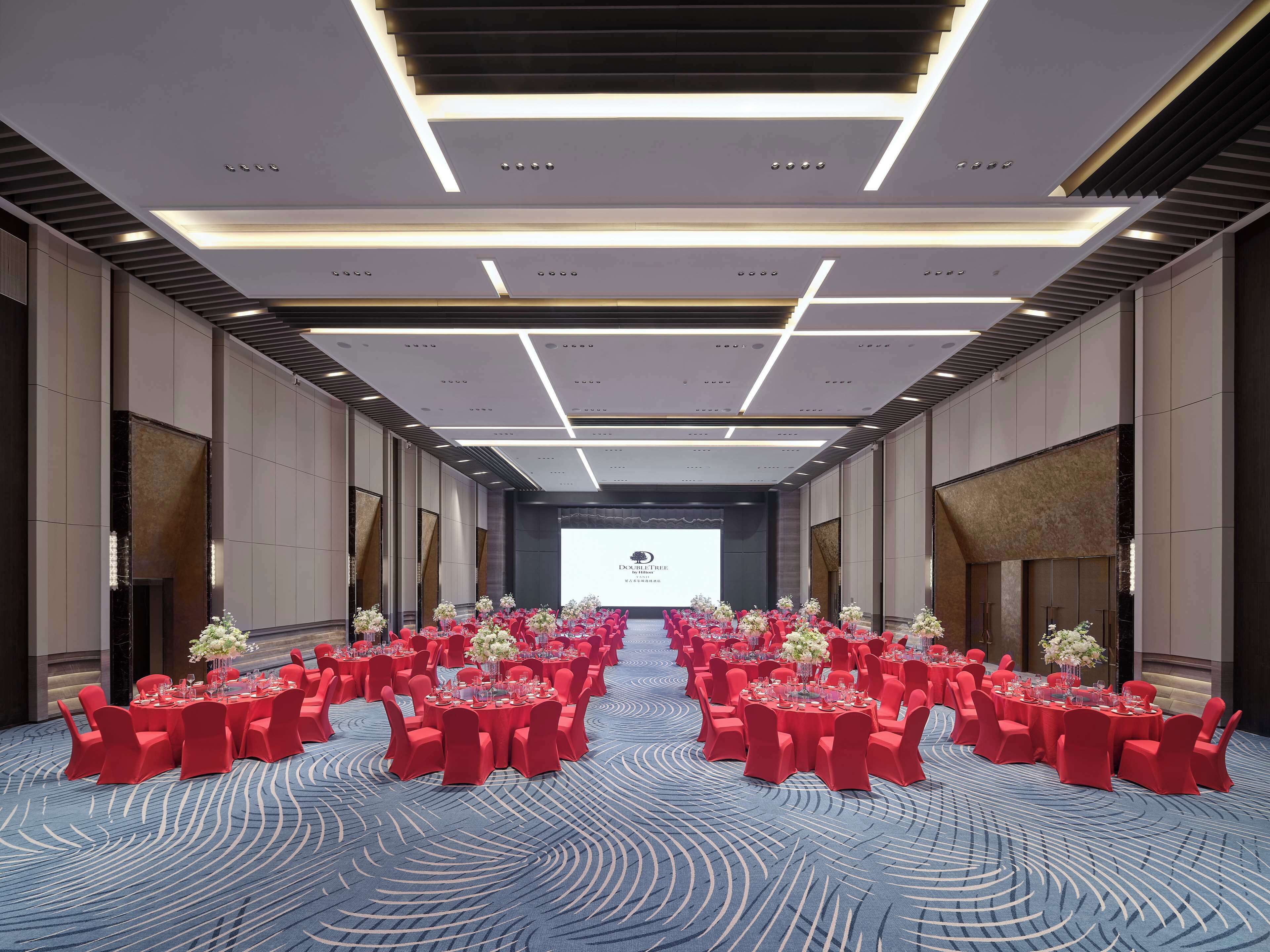 A large conference room with rows of tables and chairs, a stage at the front, and a blue carpet with a wavy pattern.