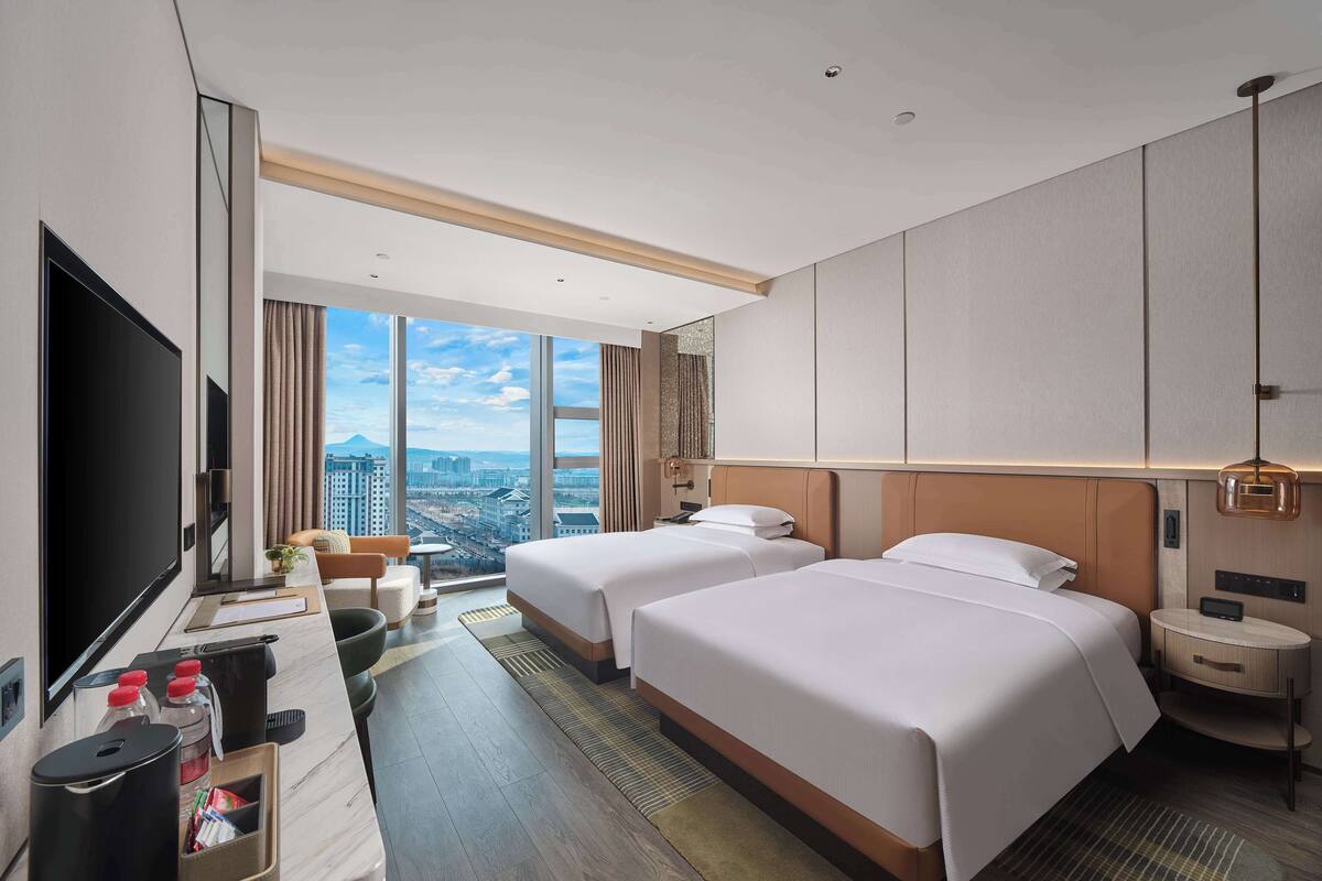 A modern hotel room with two beds, a large TV, and a city view.