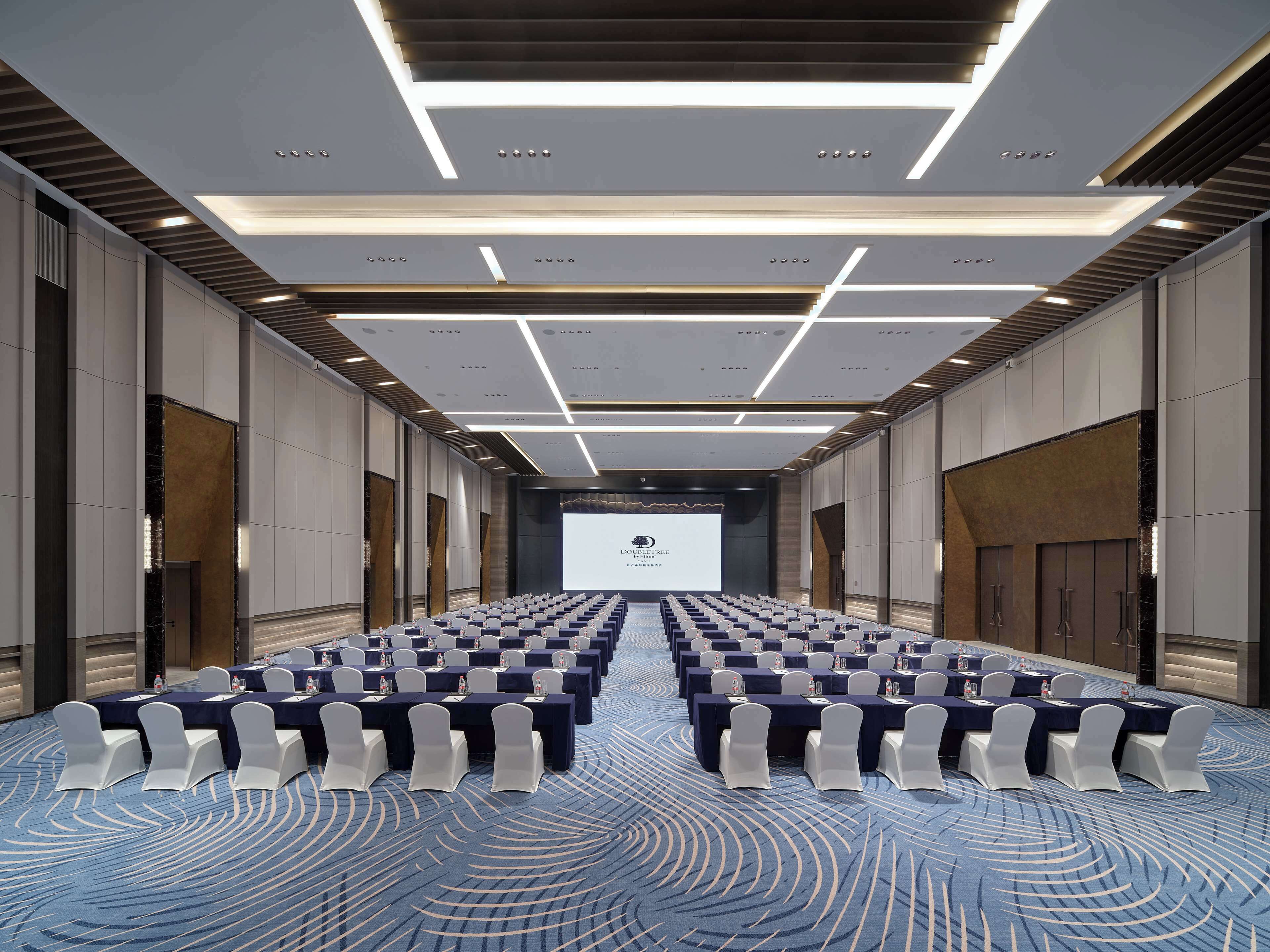 A conference room with rows of tables and chairs, a large screen at the front, and a patterned carpet.