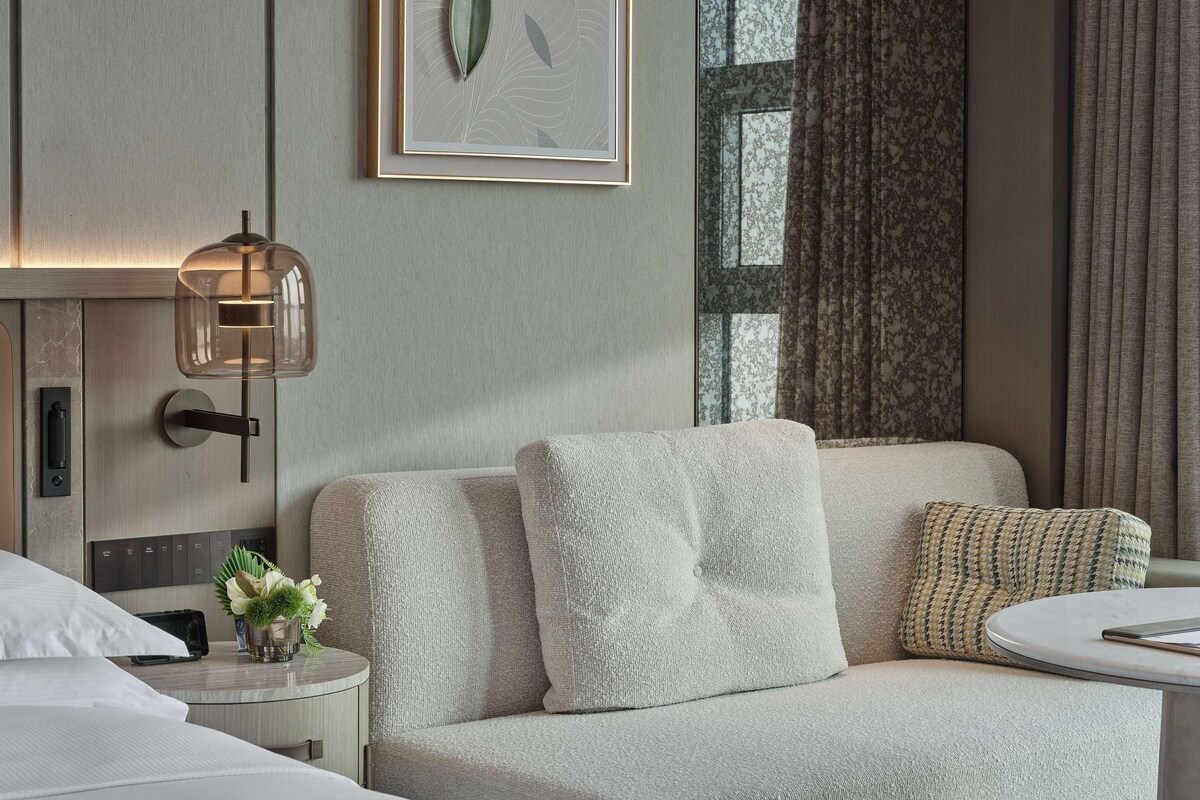 A modern hotel room with a neutral color scheme, a sofa with a textured fabric, a round side table, a wall-mounted lamp, and a framed artwork above the sofa.