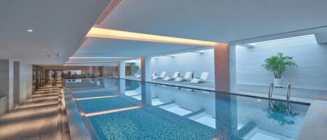 A modern indoor swimming pool with clear glass walls, a tiled floor, and a seating area with lounge chairs.