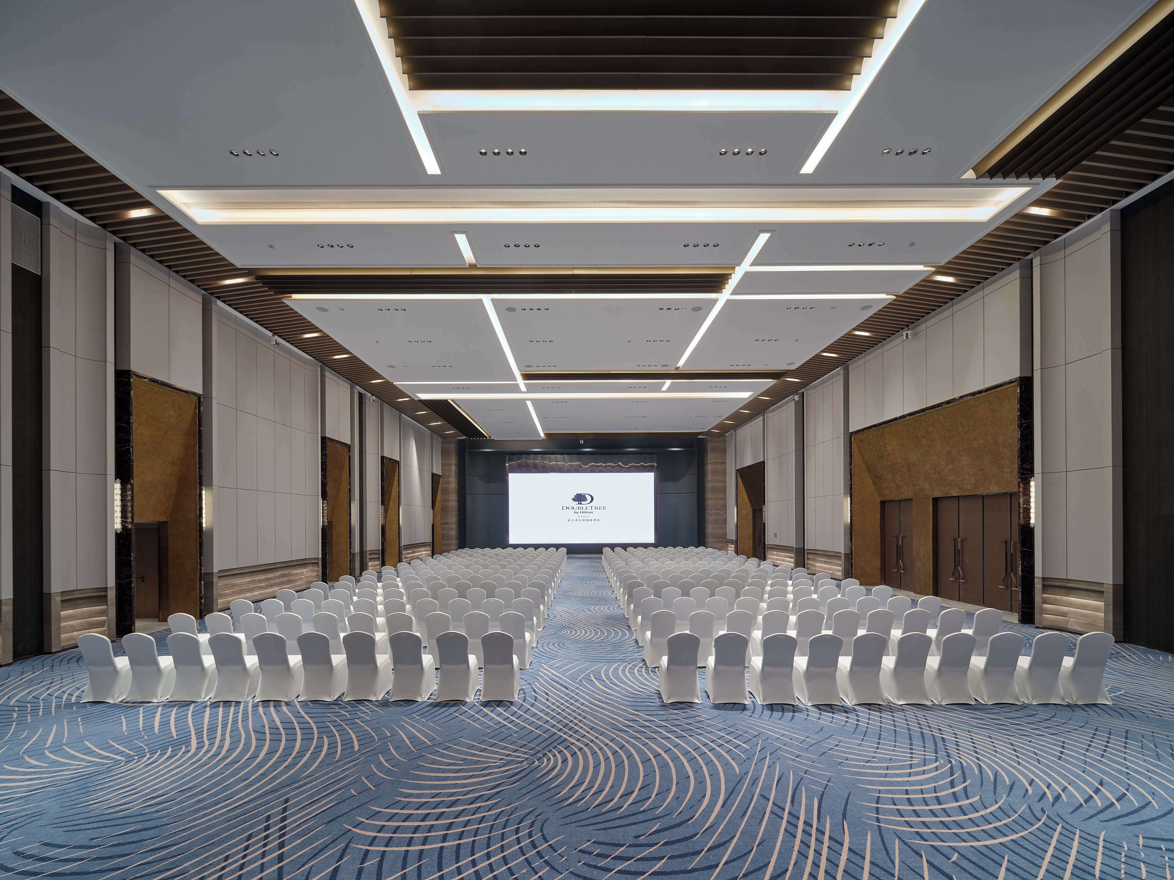 A conference room with rows of white chairs, a large screen at the front, and a patterned carpet.