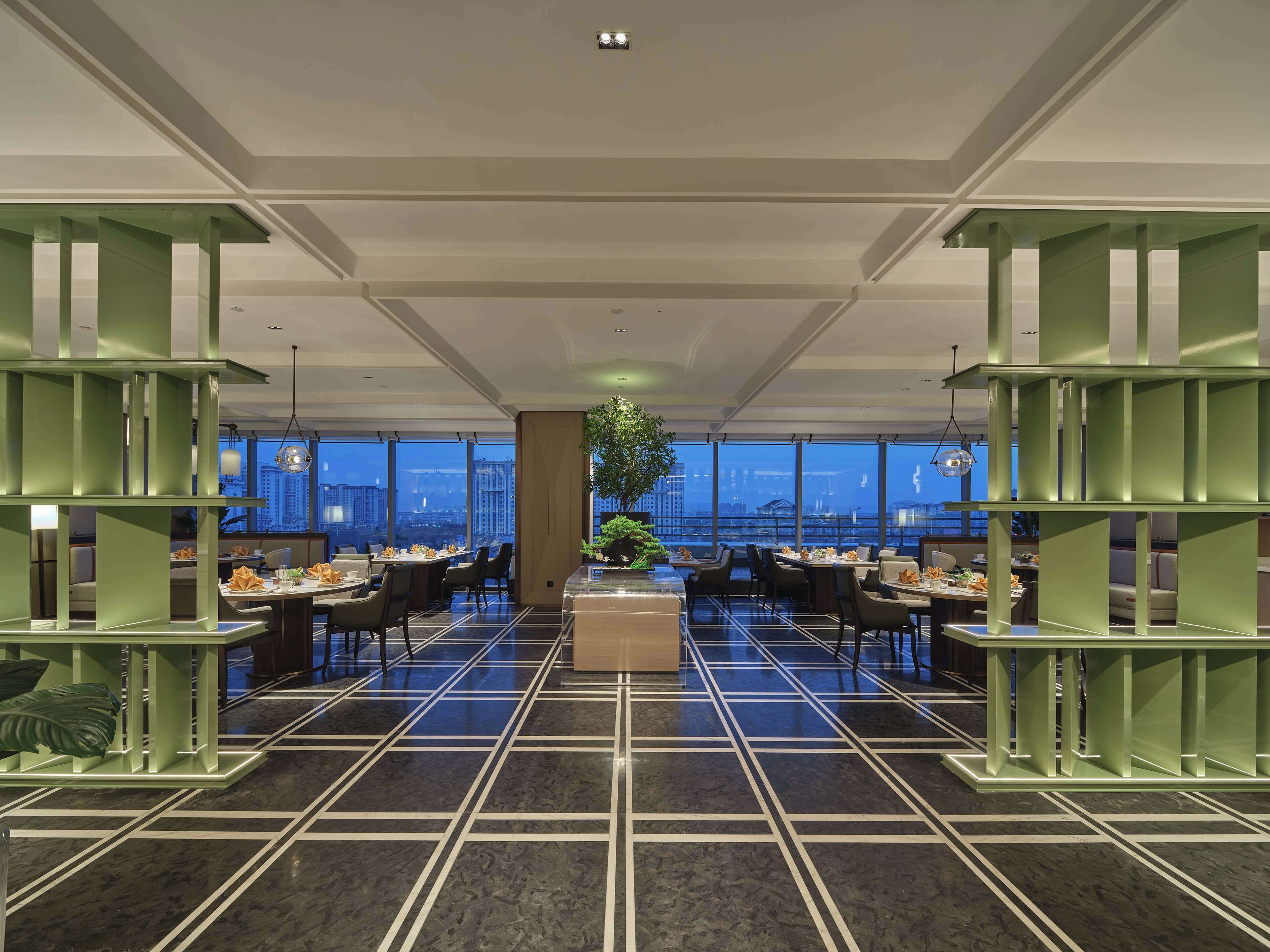 A modern dining area with a city view, featuring a central plant, green shelving, and a patterned floor.
