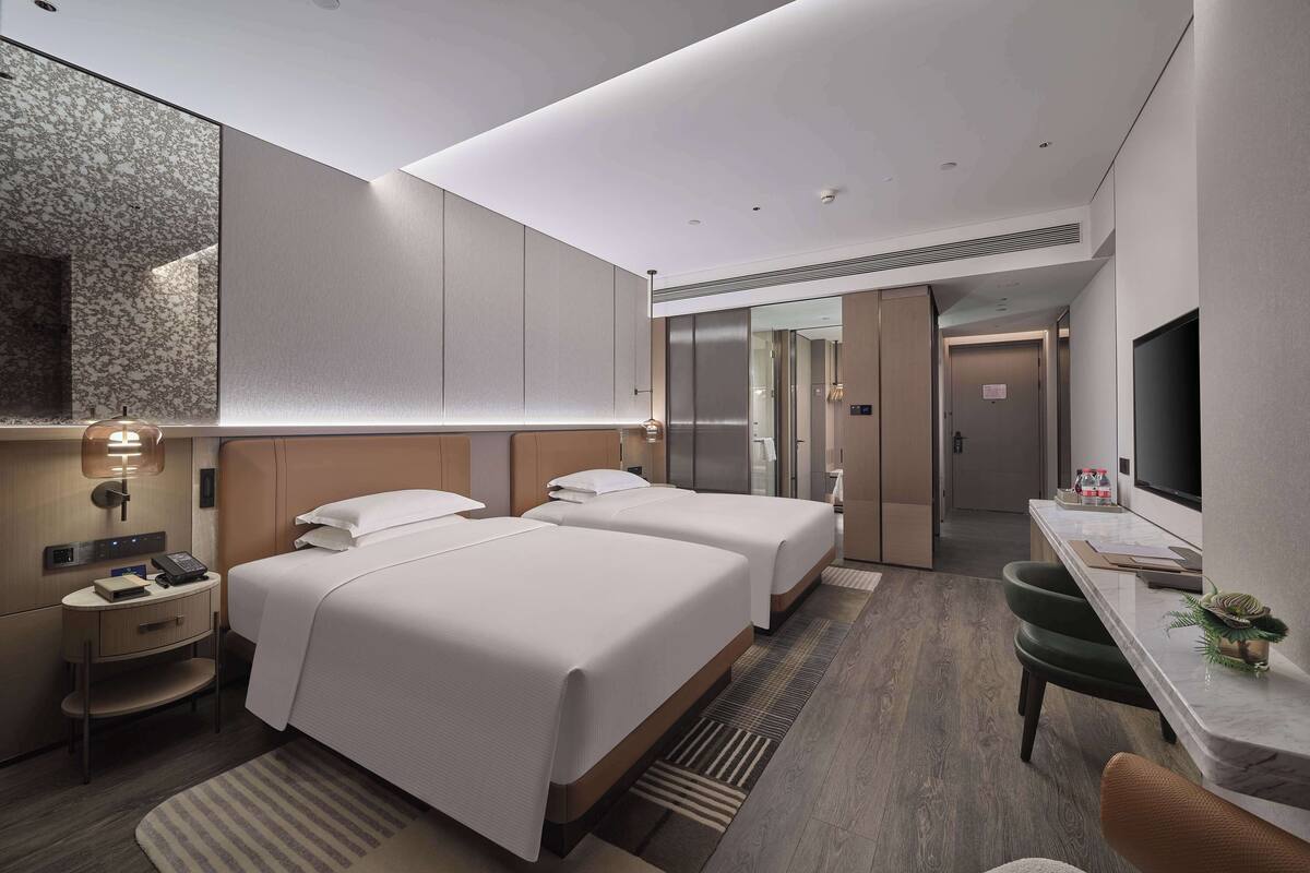 A modern hotel room with two beds, a desk, a TV, and a bathroom visible through a glass door.