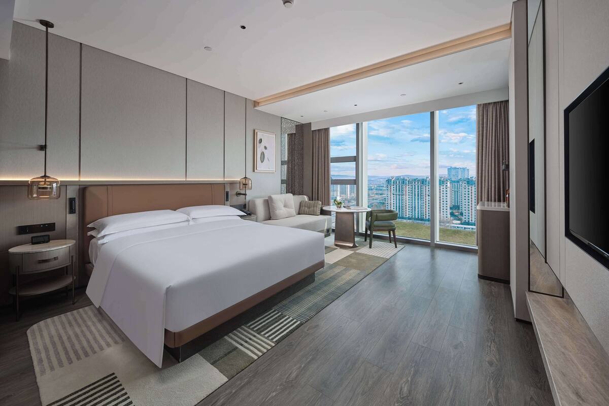 A modern hotel room with a large bed, a seating area, a dining table, and a TV.