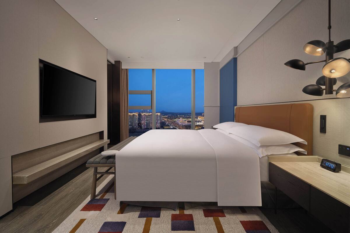 A modern hotel room with a large bed, a TV mounted on the wall, a desk with a computer, and a city view through the window.