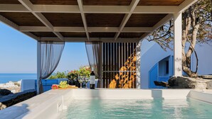 A hot tub with a view of the sea, a table with drinks, and a blue building in the background. - SunSea Living Suites (Santorini)