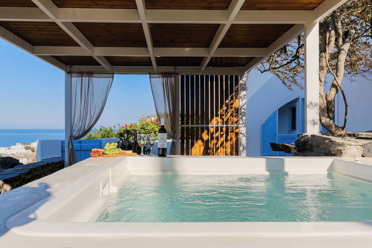A hot tub with a view of the sea, a table with drinks, and a blue building in the background.