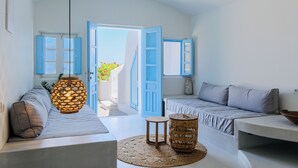 A bright, airy room with a large open door leading to a balcony, a sofa, a small table, and a wicker basket. - SunSea Living Suites (Santorini)