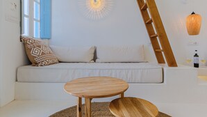 A modern living room with a white sofa, two wooden side tables, a woven rug, a wooden ladder, a wall-mounted light fixture, and a wine bottle on a table. - SunSea Living Suites (Santorini)
