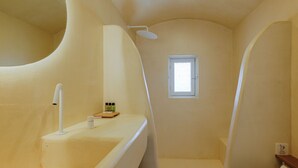 A bathroom with a curved wall, a shower, a sink, and a small window. - SunSea Living Suites (Santorini)