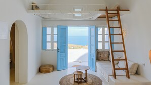 A room with a loft bed, a ladder, a small table, and a view of the beach. - SunSea Living Suites (Santorini)