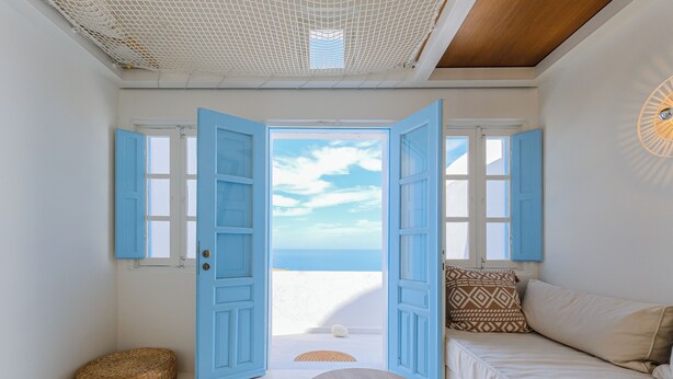 A room with a view of the sea, a wooden table, a woven stool, and a sofa with patterned cushions. - SunSea Living Suites (Santorini)