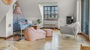Un loft spacieux avec un plafond haut, des poutres apparentes, une grande fenêtre offrant une belle vue, un canapé bleu, un fauteuil gris et un pouf rose.