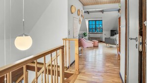 Un vaste espace de vie agrémenté d’un escalier en bois, une cuisine à l’arrière-plan et une grande fenêtre laissant entrer la lumière naturelle.