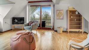 Un salon spacieux avec un plafond en bois, une télévision, un fauteuil confortable, un pouf et une bibliothèque.