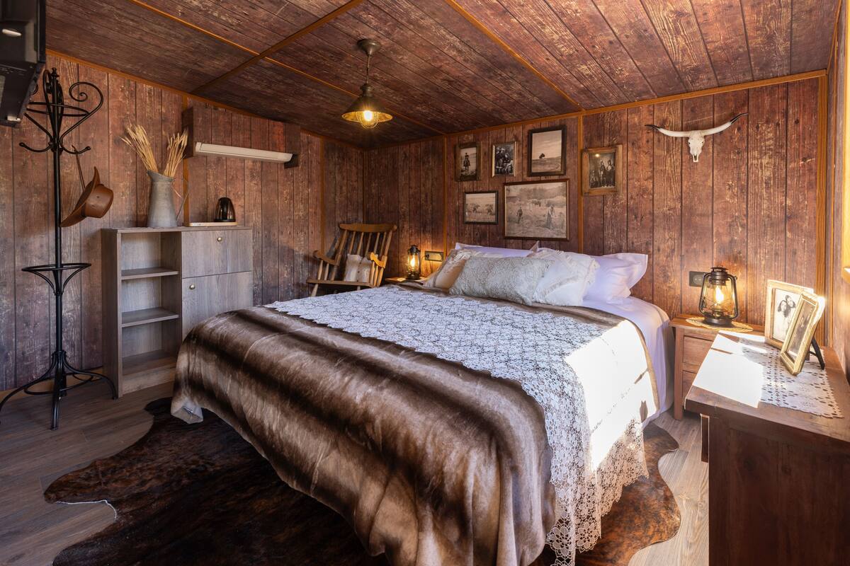 A rustic wooden room with a large bed, a wooden dresser, a chair, and a small table with a lamp.