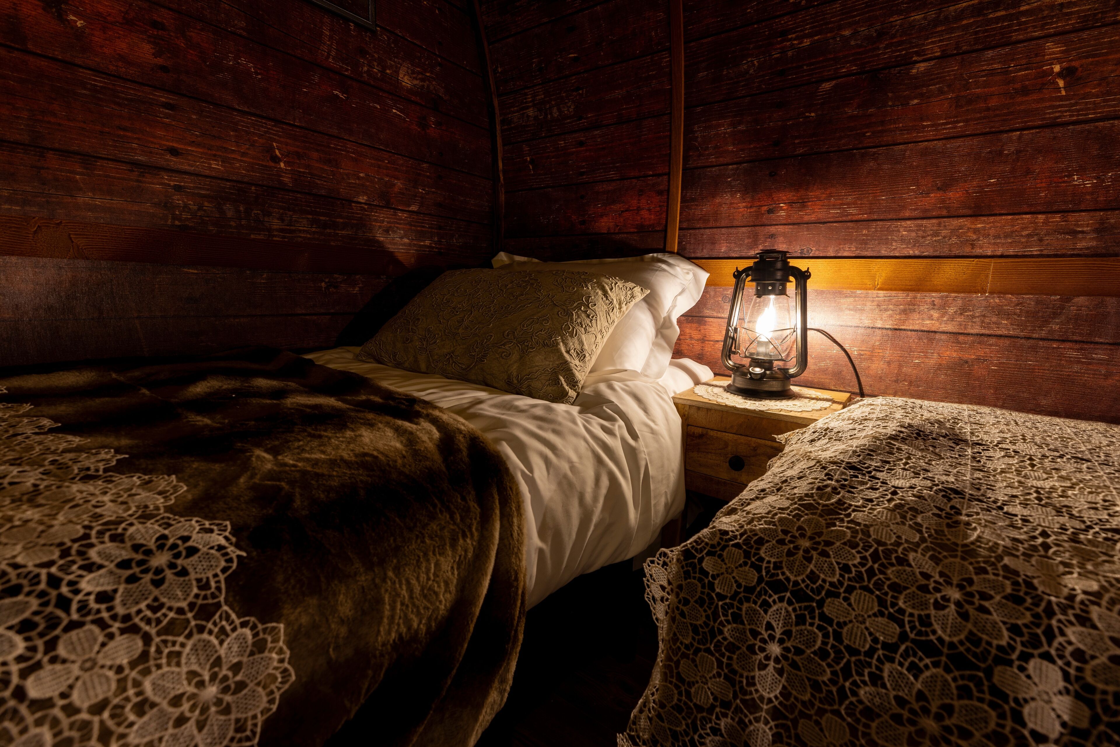 Una habitación de cabaña de madera con una cama, una mesita de noche con un farol y cubrecamas de encaje.