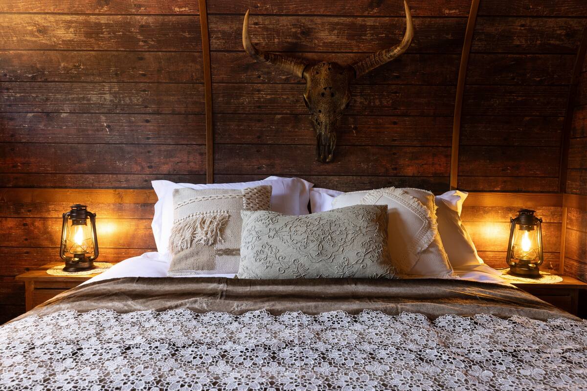 A rustic wooden room with a large bed, two bedside lanterns, and a cow skull mounted on the wall.