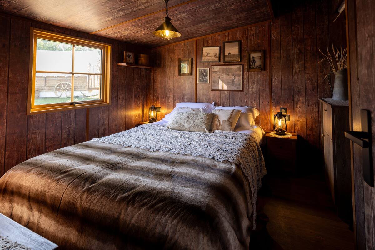 A rustic wooden room with a large bed, two bedside tables with lamps, and a window with a view of a fence and a wagon wheel.