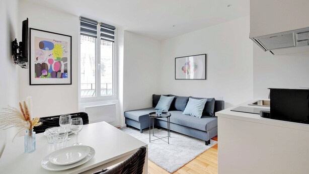 A modern living room with a grey sofa, a dining table with plates and glasses, and a kitchen area with a stove.