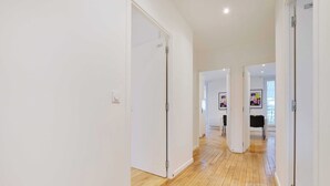 A hallway with white walls and wooden flooring, leading to a room with a black table and chairs.