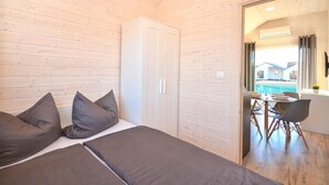A bedroom with a bed, two pillows, a wardrobe, and a view of a dining area with a table and chairs. - Holiday Home Near Bukowo Lake and Beach (Darlowo)
