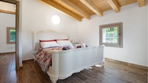 Quarto com cama branca, vigas de madeira e janela com vista para vegetação.
