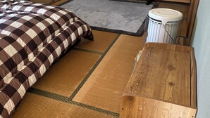 Une pièce avec un sol recouvert de tatamis, un lit avec une couverture à carreaux, une table en bois, un téléviseur posé sur un meuble et une petite poubelle blanche.