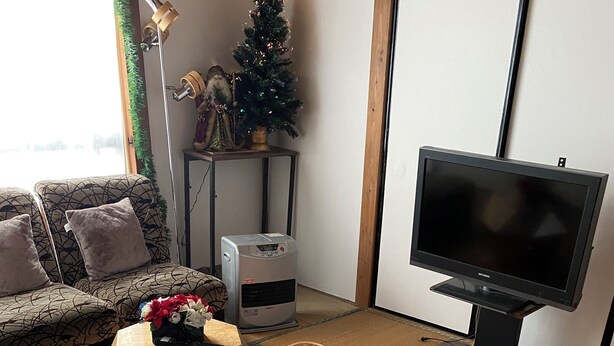 Un salon avec un sol recouvert de tatamis, un canapé agrémenté de coussins à motifs, un petit sapin de Noël, un radiateur et un téléviseur à écran plat.