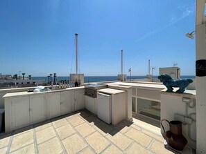 A rooftop terrace with a view of the sea, featuring a blue horse sculpture, a grill, and a row of bottles. - Mavitur Salento Apartments (Ugento)
