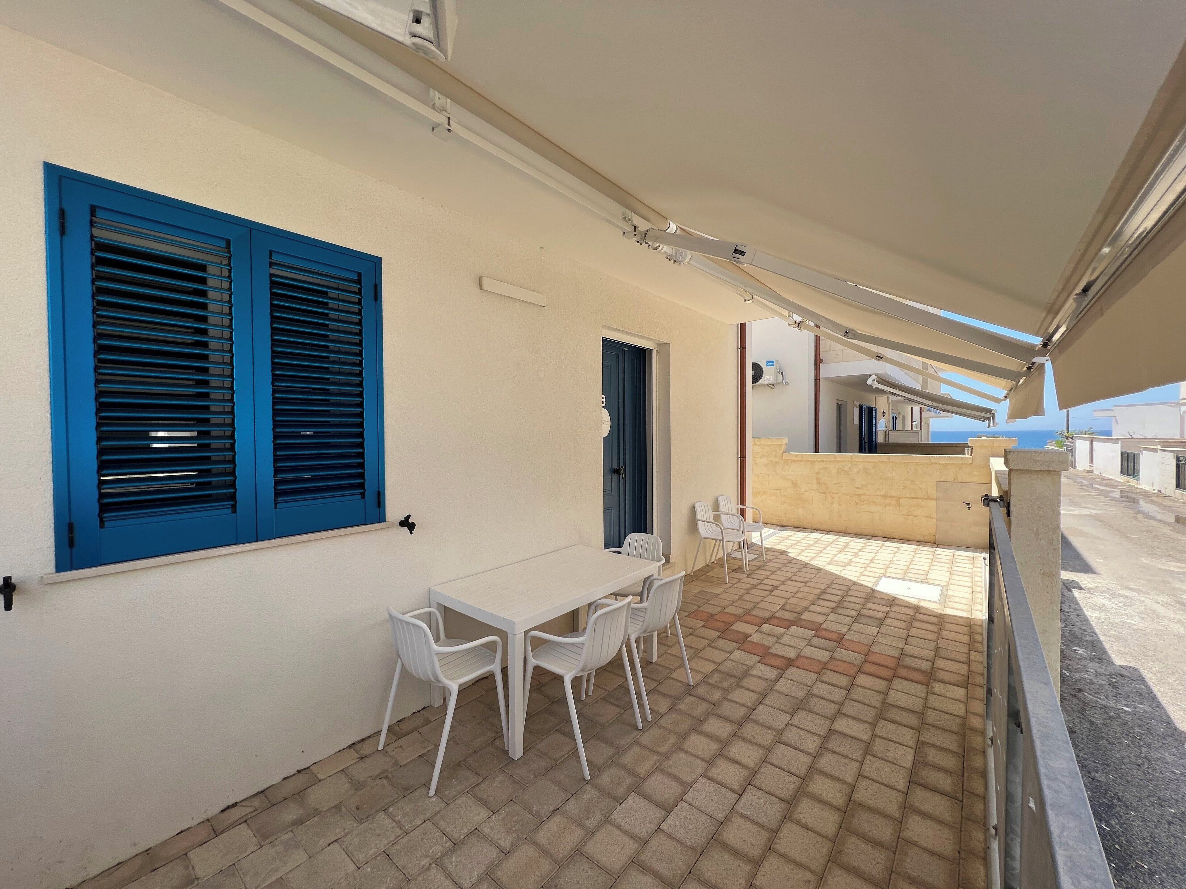 Un patio techado al aire libre con una mesa, sillas, una puerta azul y vista al mar.