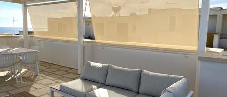 A covered outdoor seating area with a sofa and chairs, a table, and a view of the sea.