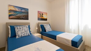 Een hotelkamer met twee bedden, elk met een blauwe sprei en passende kussens. Er hangen twee schilderijen aan de muur, voorstellende een strand en een zonsondergang.
