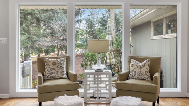 A room with two armchairs, a small table with a lamp, and a view of a garden through large windows.