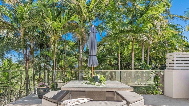 A sunlit outdoor seating area with a wicker table and chairs, surrounded by lush palm trees and a clear blue sky. - Byron Pacific Vista - Family Fun with Pool (Byron)